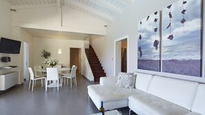 Un salon moderne avec un canapé blanc, un coin repas comprenant une table et des chaises, un escalier et un grand tableau représentant une vue sur l’océan.