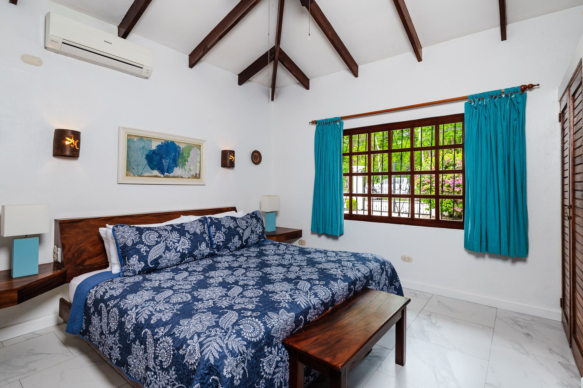 A bedroom with a bed, a bench, a window with curtains, an air conditioner, and wall-mounted lights.