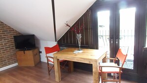 Un comedor con una mesa de madera y dos sillas rojas, un televisor sobre un mueble, y una pared de ladrillo.