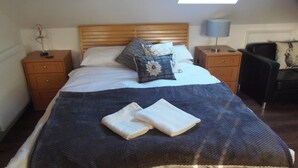 Una cama bien hecha con un edredón azul y dos toallas dobladas, cabecera de madera, mesitas de noche con lámparas y un tragaluz arriba.