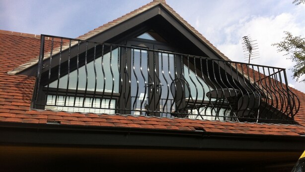 Un edificio residencial con techo inclinado, balcón con barandilla de metal y ventana con persianas.