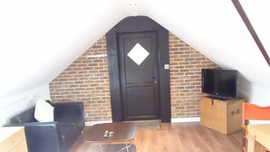 Pequeña habitación con techo inclinado, puerta negra, pared de ladrillo, un televisor sobre un pedestal, una mesa pequeña y una silla negra.