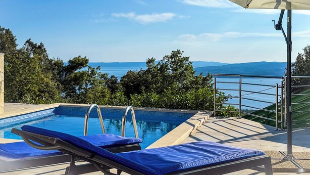 Ein Pool mit zwei Liegestühlen und Sonnensegel, mit Blick auf eine malerische Landschaft aus Meer und Bergen.