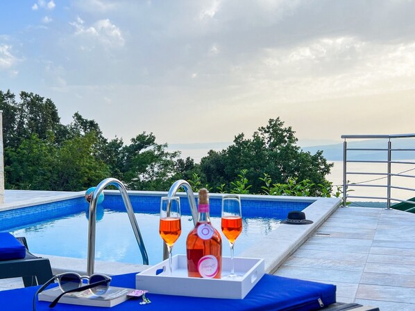 A poolside area with a view of the sea, a lounge chair, a tray with a bottle and glasses, and a metal railing.