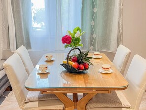 A dining table with a glass top, surrounded by white chairs, and a vase with flowers and fruit on it.