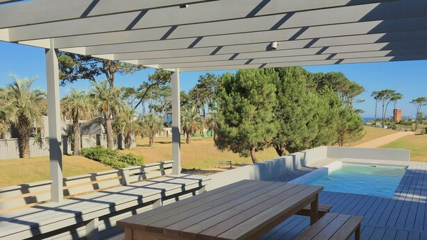A wooden deck with benches and a pool area, surrounded by trees and a clear sky.