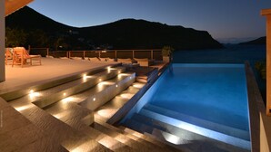 Une piscine accessible par des marches, entourée d’une terrasse en bois et offrant une vue sur la mer et les montagnes.