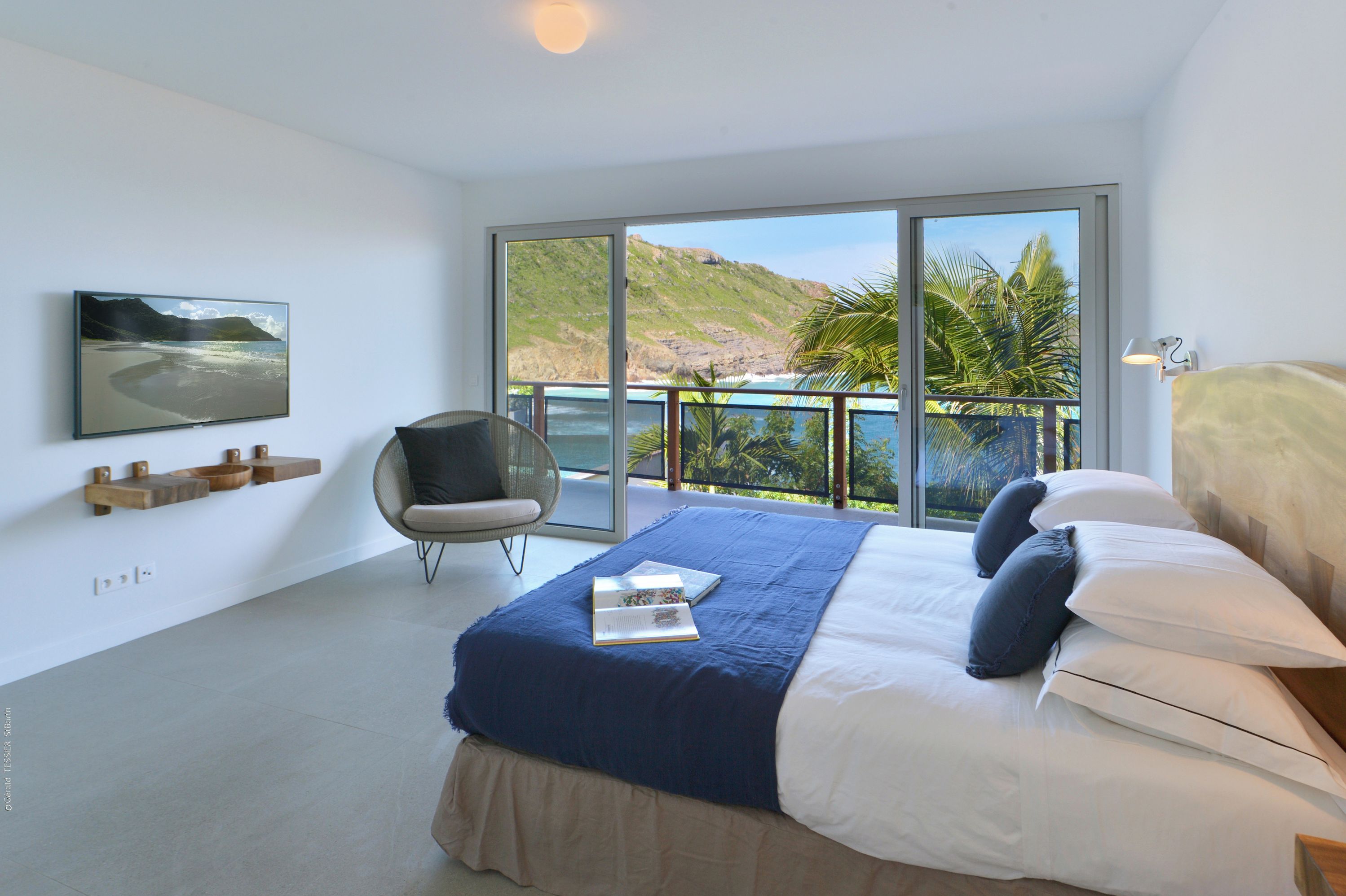 A modern bedroom with a large bed, a chair, a mounted TV, and a balcony with a view of the beach and hills.