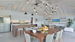Une cuisine spacieuse équipée d’appareils modernes, un coin repas avec une table en bois et des chaises en osier, et un salon doté d’une grande fenêtre donnant sur la plage.