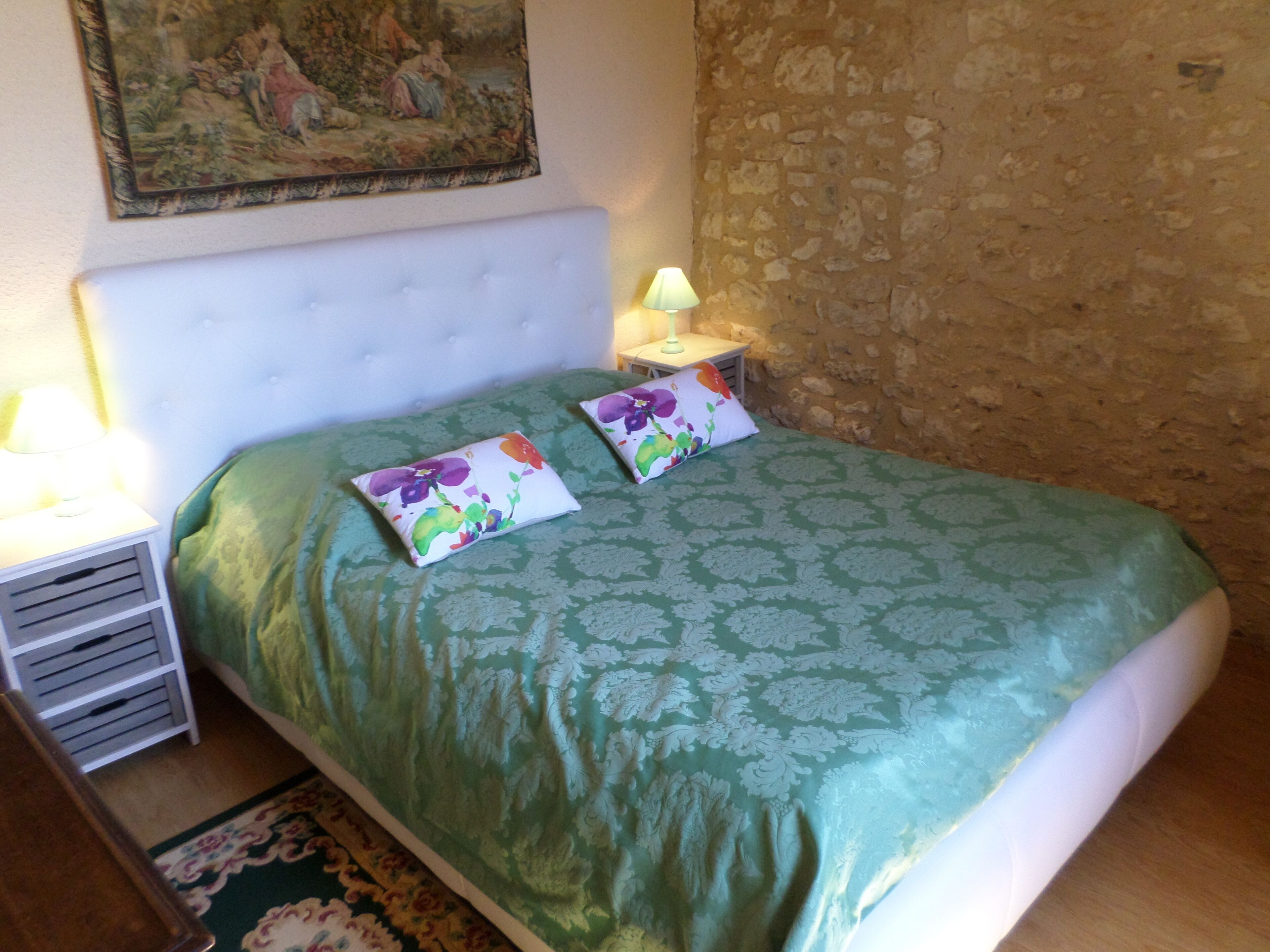 A bedroom with a green patterned bedspread, two pillows with floral designs, a white headboard, a nightstand with a lamp, and a painting on the wall.