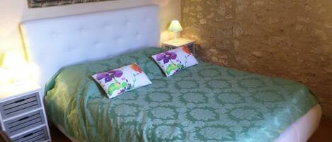 A bedroom with a green patterned bedspread, two pillows with floral designs, a white headboard, a nightstand with a lamp, and a painting on the wall.