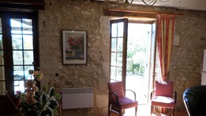 A rustic room with stone walls, a wooden ceiling, and a large window with curtains. There is a chair, a table with a vase of flowers, and a radiator. - The Country House Mijotiere (monestier)