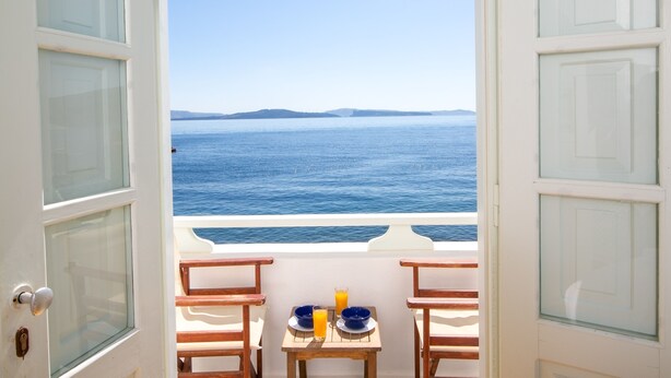 Un balcon ouvert avec une table et des chaises, donnant sur une mer calme et une terre lointaine.