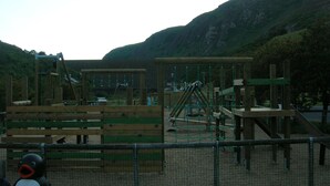 A playground with various play structures, surrounded by a fence and set against a backdrop of hills.
