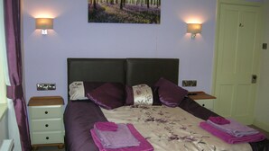 A neatly made bed with purple bedding, two bedside lamps, and a painting of a forest on the wall.