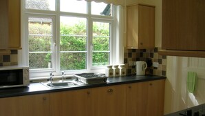 A kitchen with wooden cabinets, a microwave, a kettle, and a window with a view of greenery.