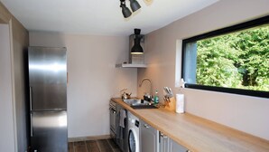 Cozinha compacta com frigorífico em aço inoxidável, máquina de lavar, pia e janela com vista para árvores.