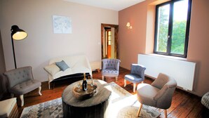Uma sala de estar aconchegante com sofá, poltronas, uma mesa de centro com garrafa e copos, um abajur e uma janela com vista para o verde.