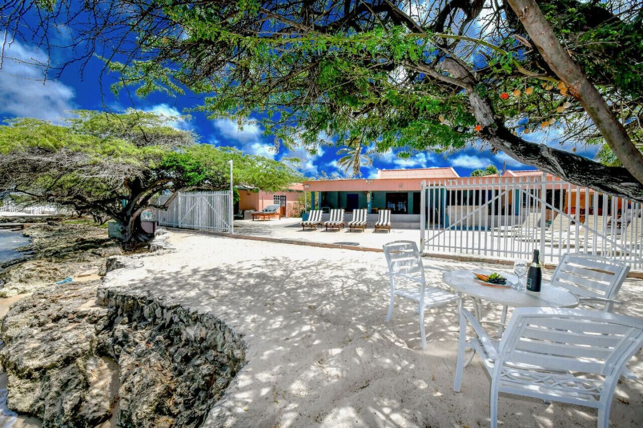 A sandy outdoor area with white lounge chairs and a table, a building with a red roof, and a tree with green leaves.
