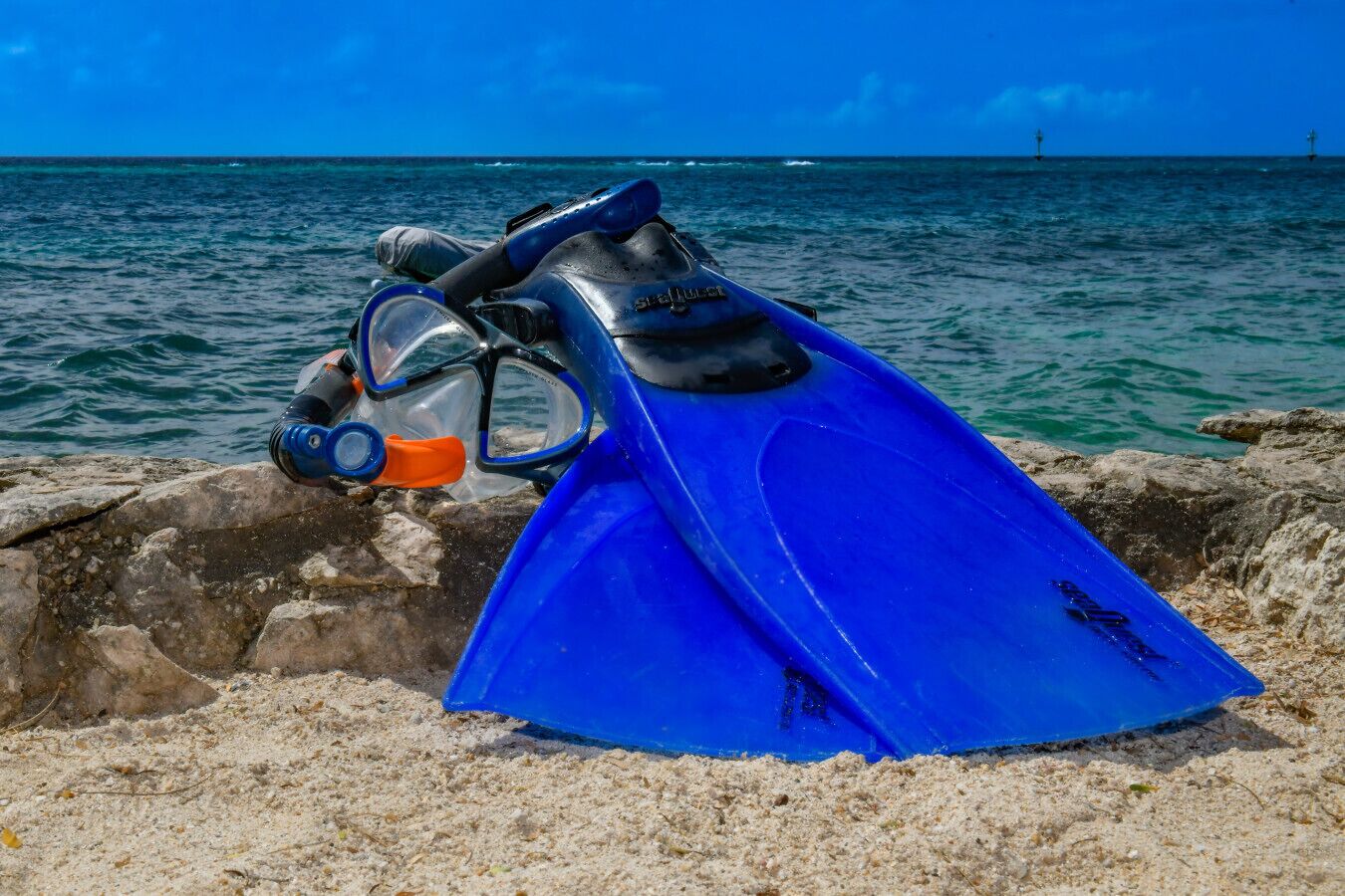A blue diving board with fins and a mask resting on a rocky surface near the ocean.