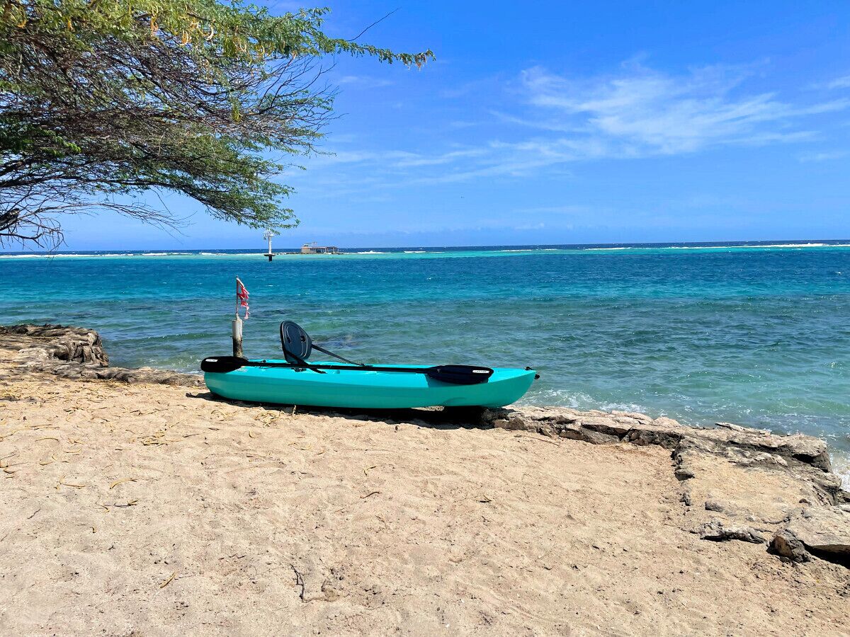 Un kayak turquesa sobre una playa arenosa con agua azul cristalina y un faro distante.