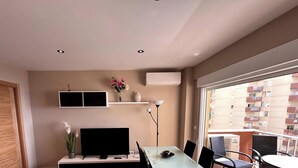 A compact living space with a dining area, a flat-screen TV, a small plant, and a window overlooking a residential building.