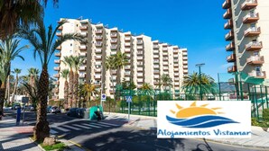 A high-rise residential building with balconies, palm trees, and a clear blue sky.