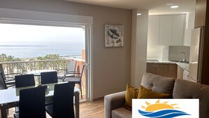 A modern living room with a sofa, a dining table with chairs, and a view of the sea.