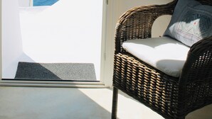 Uma cadeira de vime com almofada e travesseiro, colocada ao lado de uma parede branca com uma porta que dá para uma varanda.