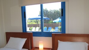 A bedroom with two beds, a nightstand with a lamp, and a window overlooking a pool area.
