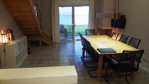 Un coin repas avec une table et des chaises en bois, un téléviseur fixé au mur et une vue sur la mer par une porte vitrée.