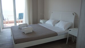 Un lit bien fait, avec du linge de lit blanc, une table de chevet avec une lampe, et une vue sur la mer par une porte vitrée.