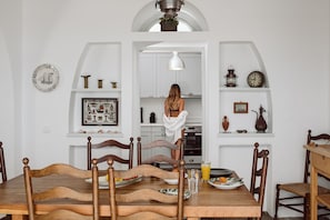 A dining area with a wooden table and chairs, a kitchen with white cabinets, and various kitchen items on shelves. - Armeni Seaside Villa - with private boat service! (Santorini)