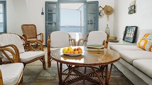 Une pièce meublée de meubles en osier, une table en verre sur laquelle repose un panier de fruits, et une vue dégagée grâce à des portes ouvertes.