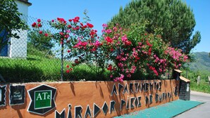Une enseigne indiquant « APARTMENTES MIRADOR PISO DE EUROPA », avec un logo « ATR » vert, entourée de fleurs roses en pleine floraison et d’une clôture.