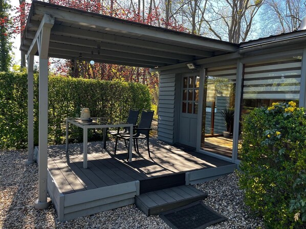 A covered patio with a table and chairs, a small step leading to a door, and a garden with a hedge and yellow flowers.