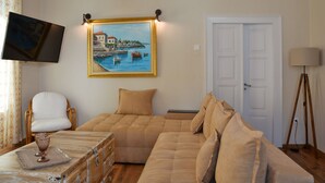 Een woonkamer met een beige bank, een witte fauteuil, een flatscreen-televisie, een schilderij van een haven en een lamp.