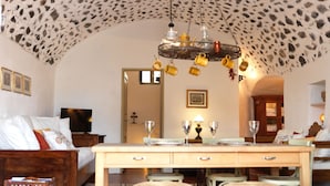 Een kamer met een stenen plafond, een houten eettafel met stoelen, een bed met wit beddengoed en een televisie op een houten dressoir.