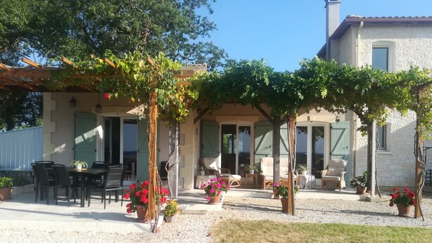 Een overdekt terras met tafels en stoelen, omgeven door plantenpotten en een pergola met klimplanten.