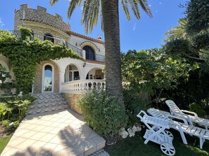 A stone building with arched windows and a balcony, surrounded by lush greenery and palm trees.