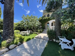 A garden with a paved walkway, white outdoor furniture, and a stone building with arches.
