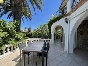A covered outdoor patio with a dining table and chairs, surrounded by white columns and arches, with a view of a clear blue sky and palm trees.