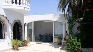 Uma área de piscina com um edifício branco, um alpendre coberto com cadeiras e plantas em vasos.
