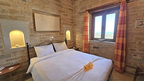 Une chambre avec un mur en pierre, un lit recouvert de draps blancs, deux lampes fixées au mur et une fenêtre avec des rideaux.