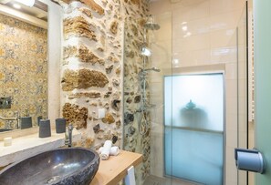 A bathroom with a stone wall, a large sink, a glass shower enclosure, and a mirror. - Timeless Elegance Embraced by Comfort and Bliss (Rethymno)