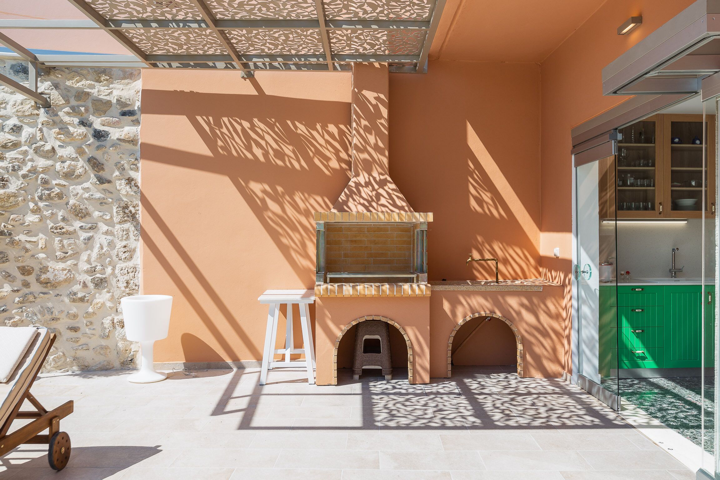 Une terrasse avec un mur en pierre, un tabouret blanc, une chaise en bois, une petite maison pour chat et un meuble vert.