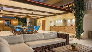 Amplia zona de patio con mesa de comedor, sillas azules, un sofá de mimbre y una cocina al fondo.
