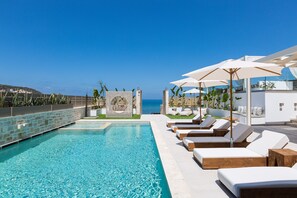 A poolside area with lounge chairs and umbrellas, a clear view of the sea, and a stone wall with a decorative pattern. - Villa Moana - Beachfront Coastal Dreams by etouri (Rethymno)