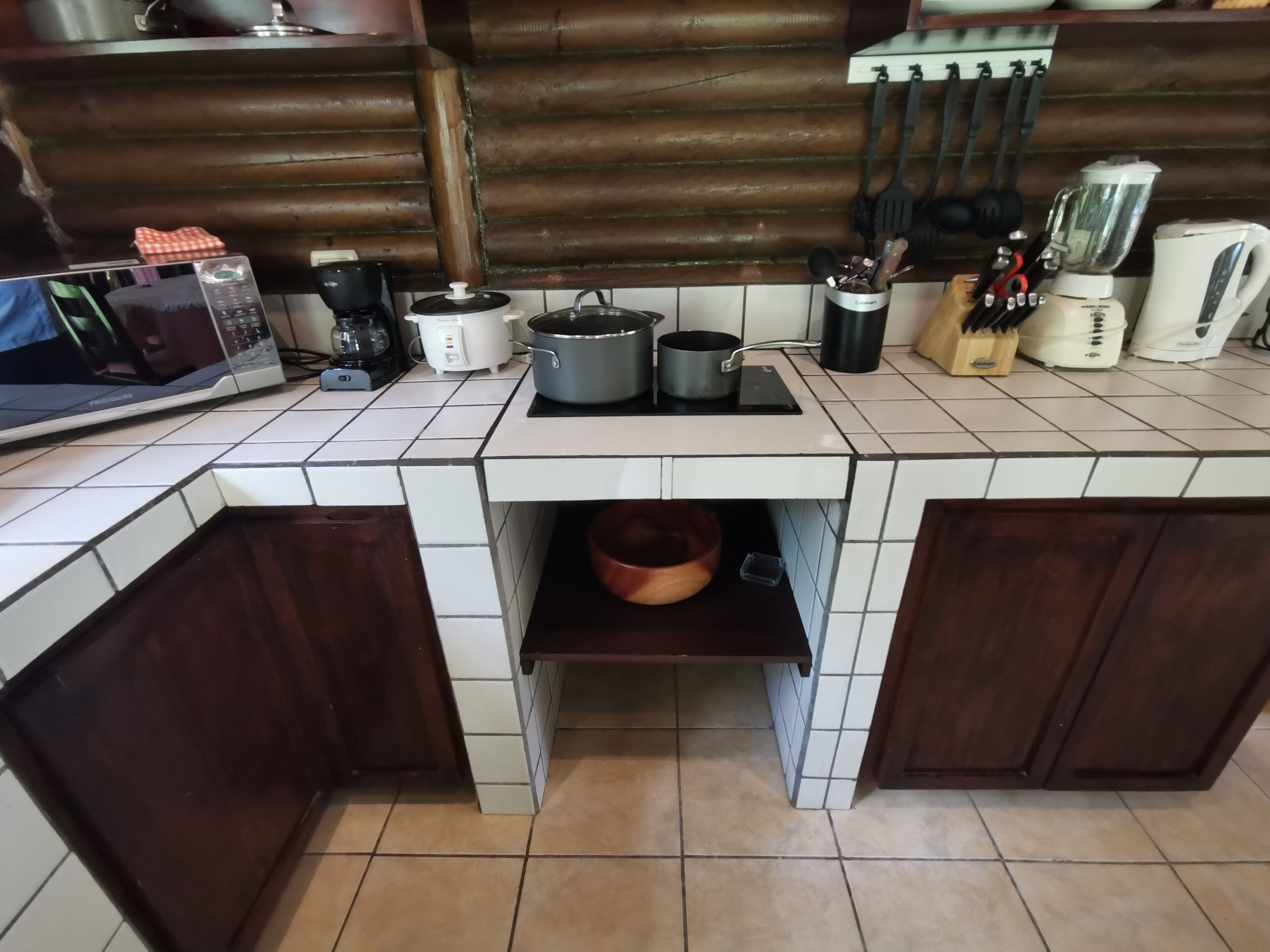 A kitchen with a tiled backsplash, wooden cabinets, a countertop with various appliances, and a hanging utensil rack.