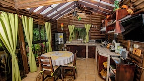 A rustic wooden cabin with a dining area featuring a round table and chairs, a kitchen with a refrigerator and a microwave, and a ceiling fan. - Cabaña Los Tucanes - Paradise for Couples! (La Fortuna, Costa Rica)